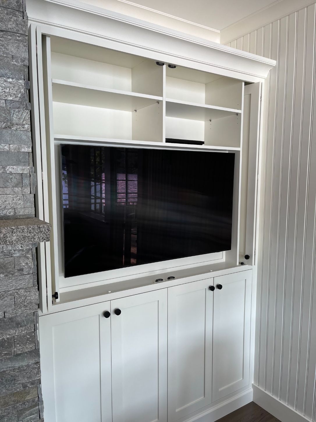 White built-in media wall with a recessed TV, open shelves above and cabinets below, beside a stone fireplace