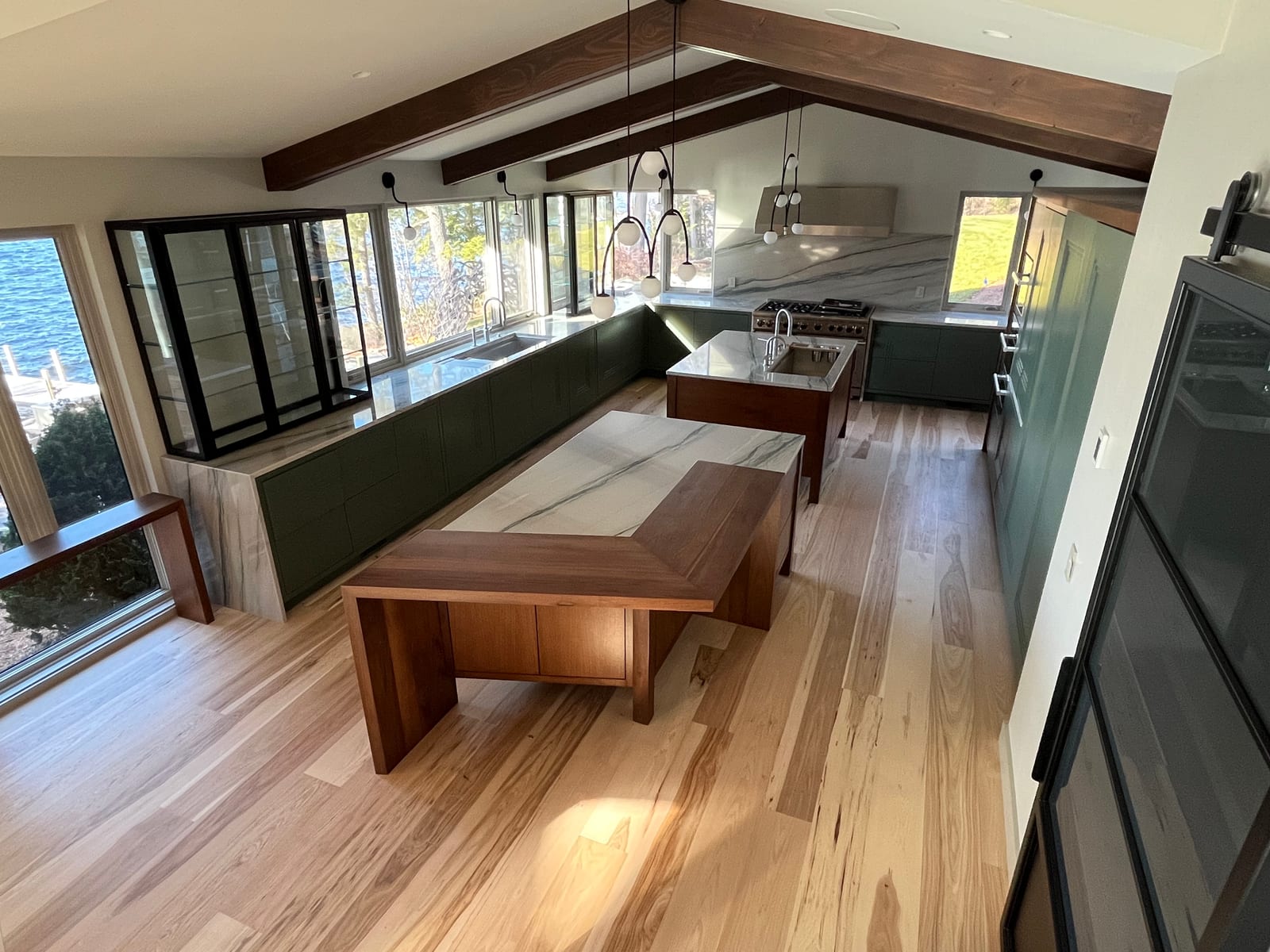 Modern lakeside kitchen with deep green flat-panel cabinetry, a book-matched marble backsplash and waterfall island, walnut ceiling beams and a hickory floor, built by NH Woodworks